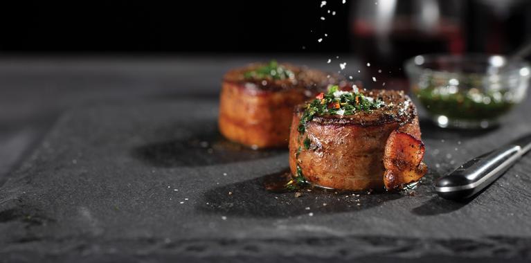 Bacon-wrapped filet mignon, seasoned and grilled on a gray platter with garnish